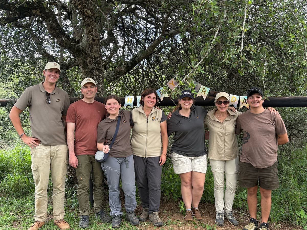 A group of wildlife conservation volunteers and staff standing together outdoors under a tree, smiling in front of a “Happy Birthday” banner in a bush setting.