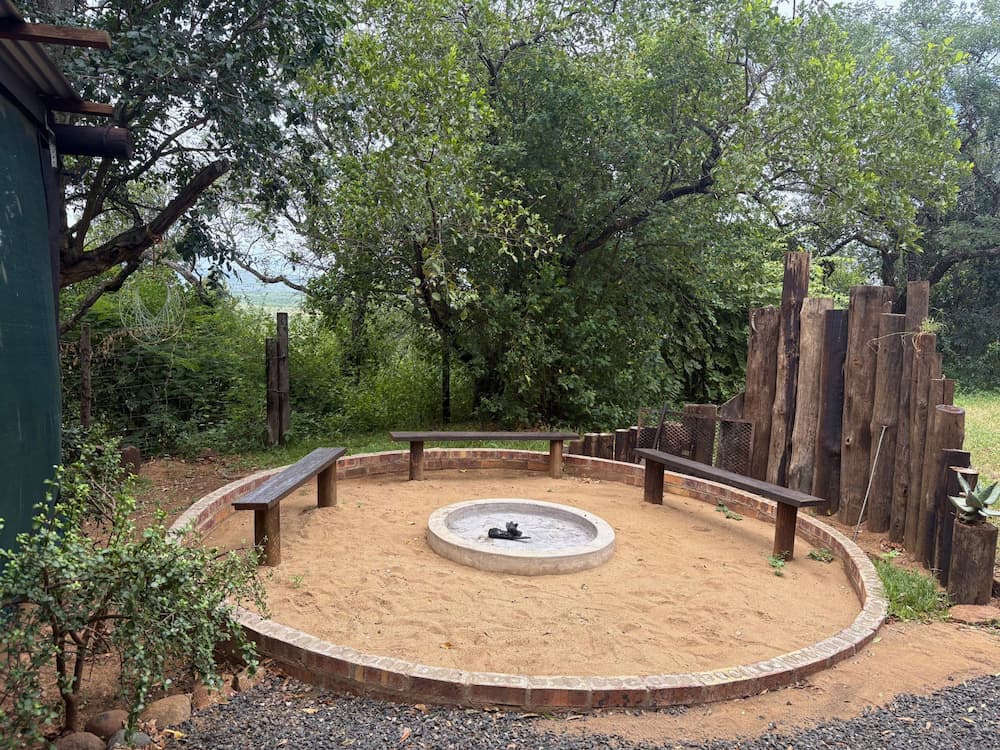 A circular outdoor seating area with wooden benches arranged around a central fire pit, set in a shaded, leafy bush environment near a conservation camp.