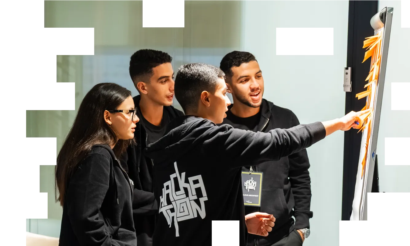 Four young people in black hoodies discussing and pointing at sticky notes on a board in a bright room.