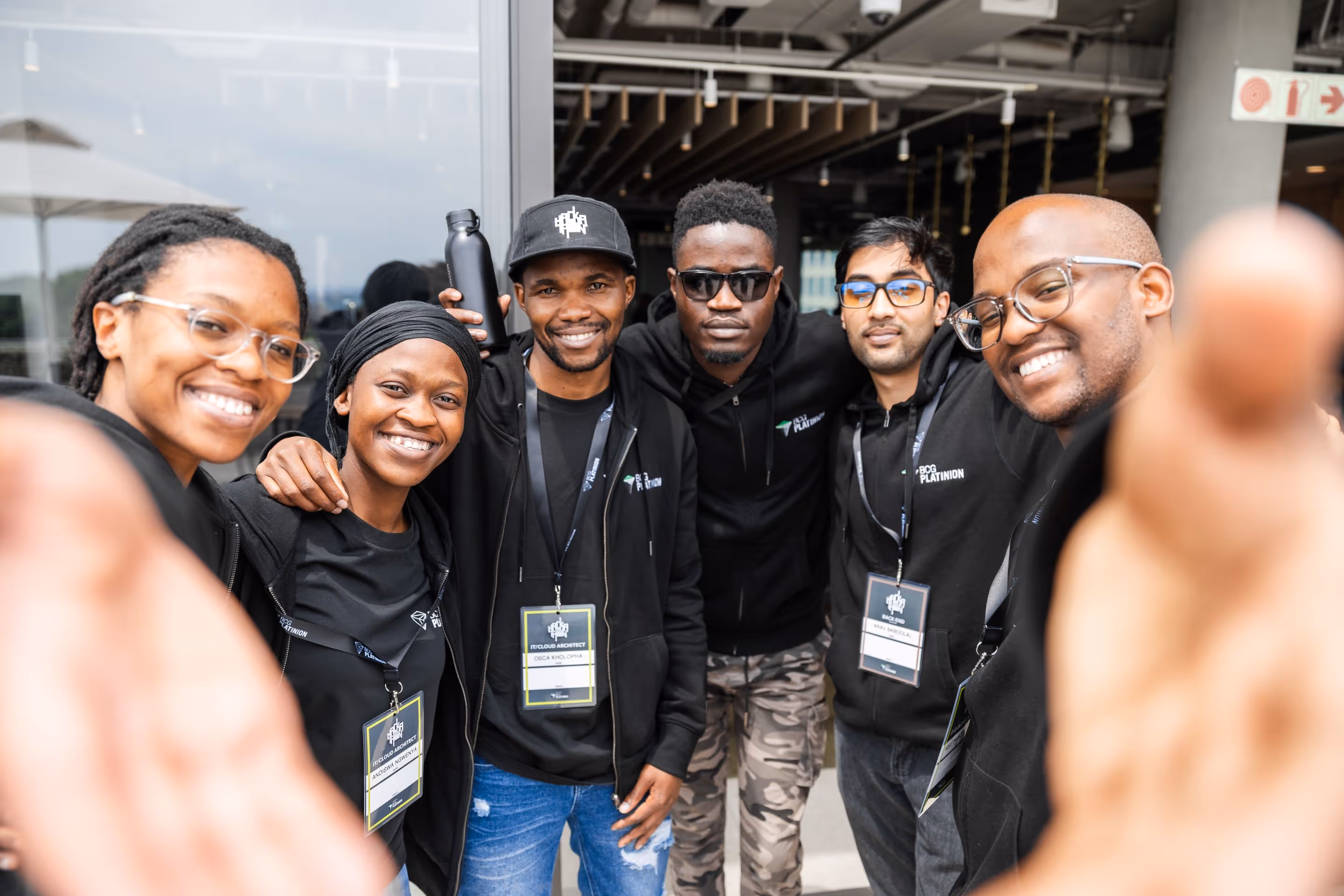 Six smiling diverse young adults in black hoodies with badges reaching toward the camera.