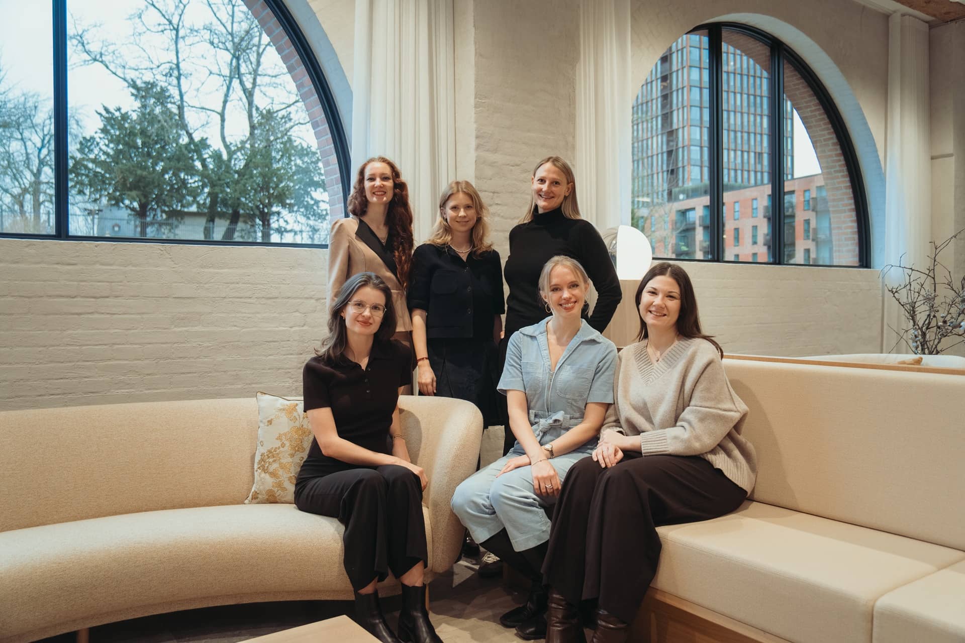 Group of six smiling women seated and standing in a modern room with arched windows and beige couches.
