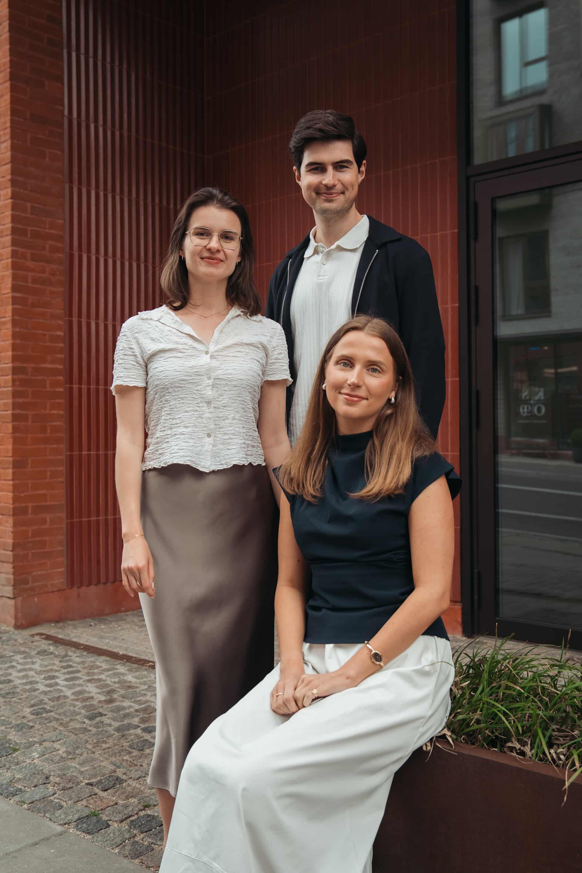 Three young adults posing outdoors by a building; one seated, two standing, all smiling at the camera.