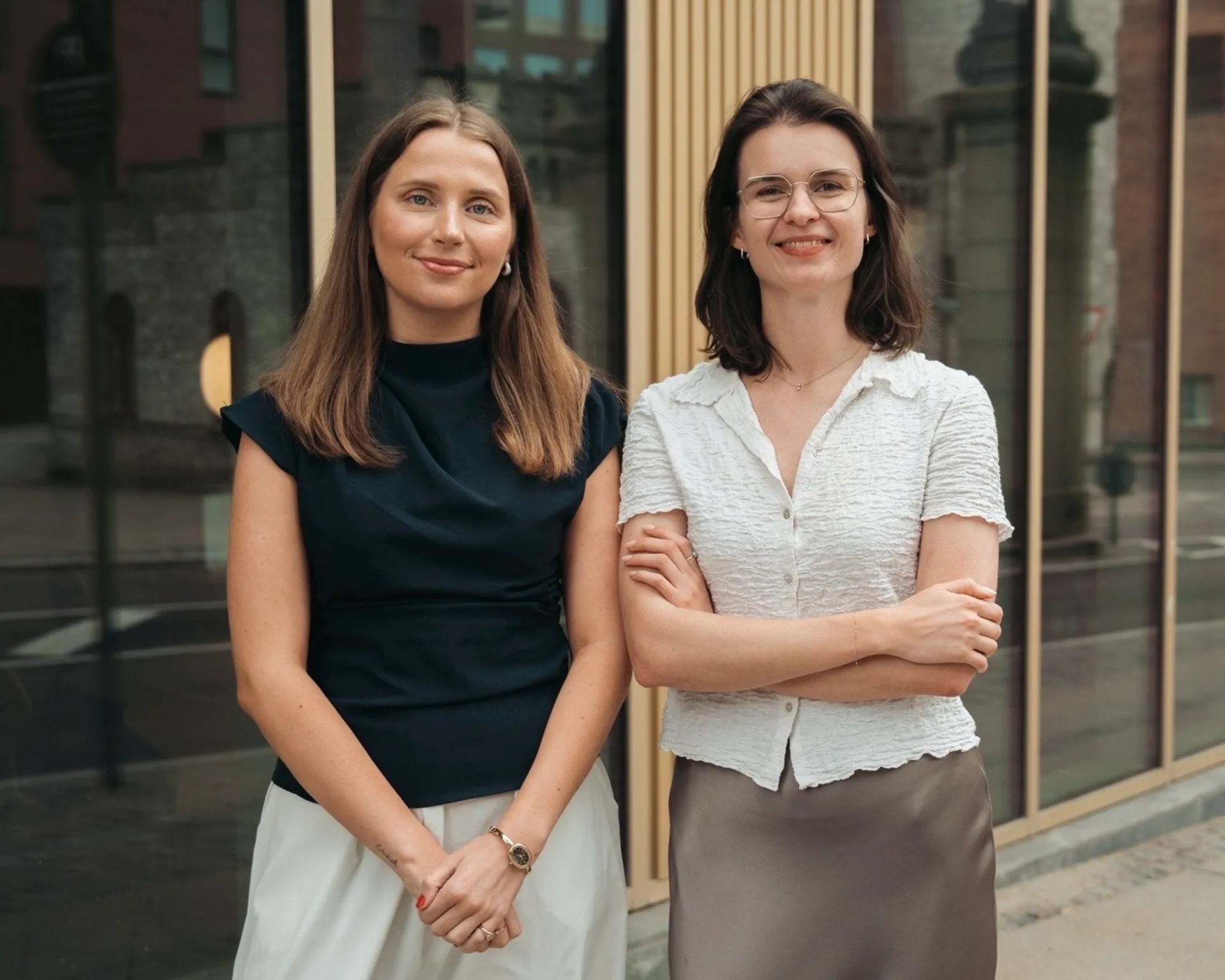 Two women stand side by side smiling; one wears black top and white skirt, the other wears white shirt and gray skirt.