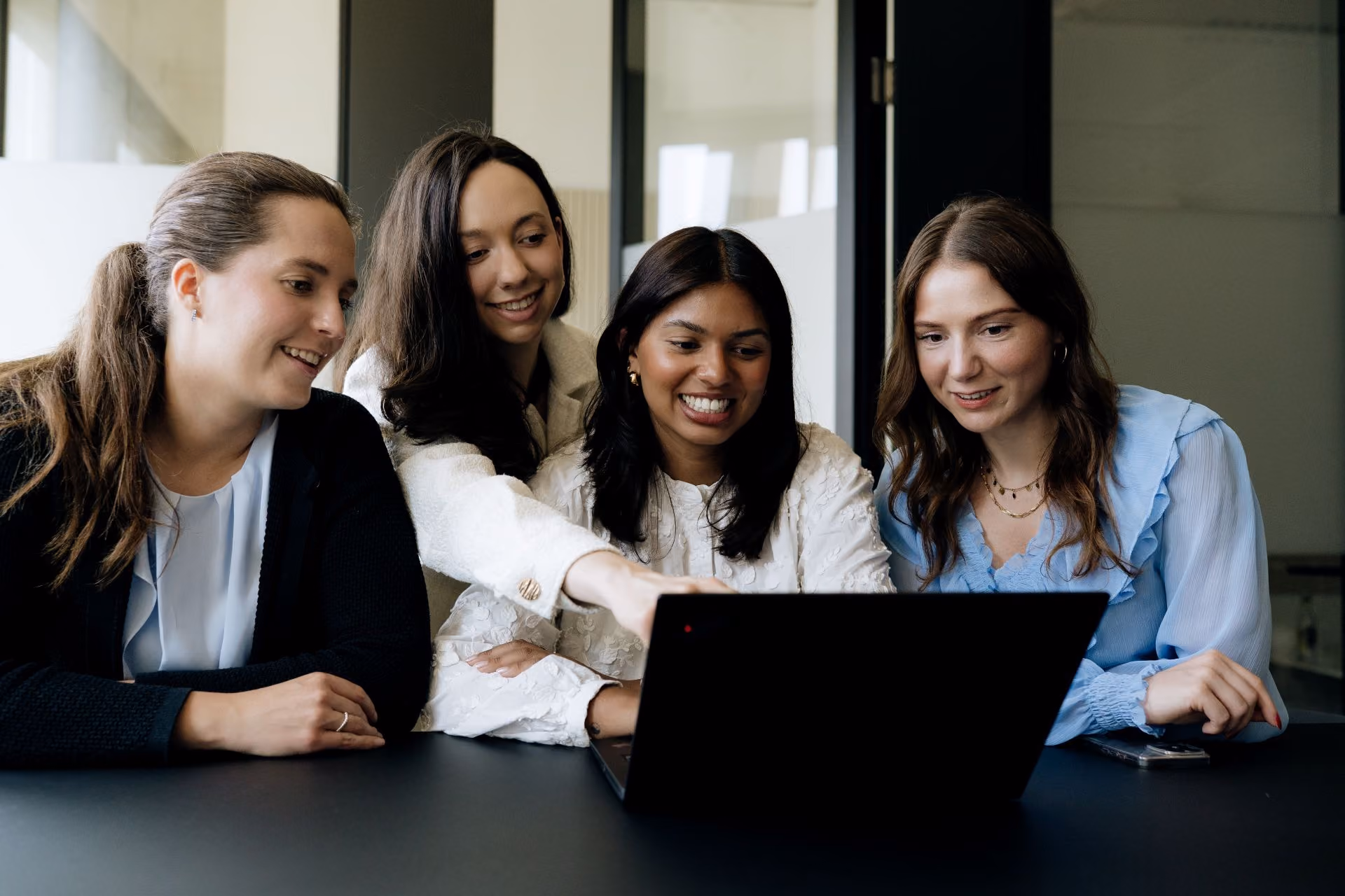Four women sitting at a table, smiling and looking at a laptop in front of them, with one woman pointing at the screen.