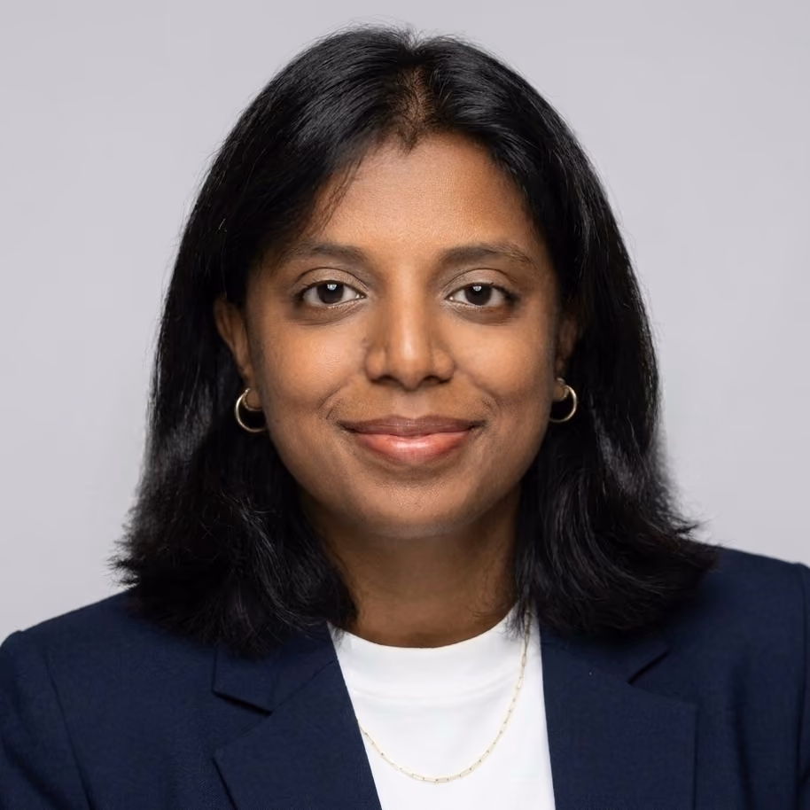 Portrait of a woman with medium-length black hair wearing a navy blazer, white top, small gold hoop earrings, and a delicate gold chain necklace, smiling softly against a plain light gray background.