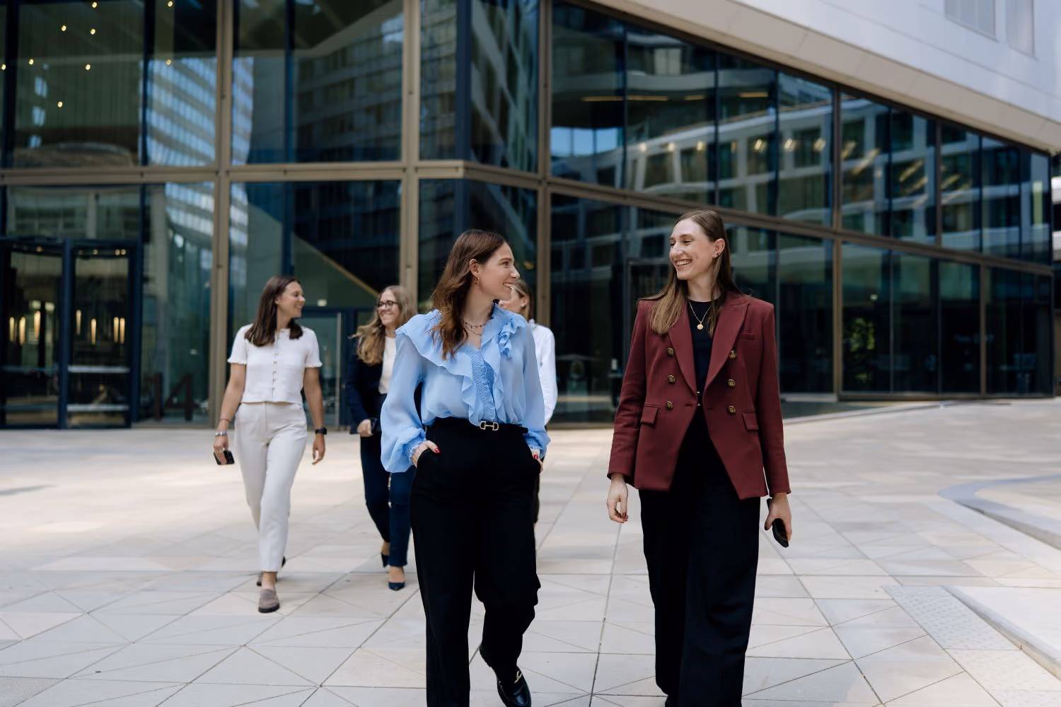 Five professionally dressed women walking and talking outside a modern glass office building.