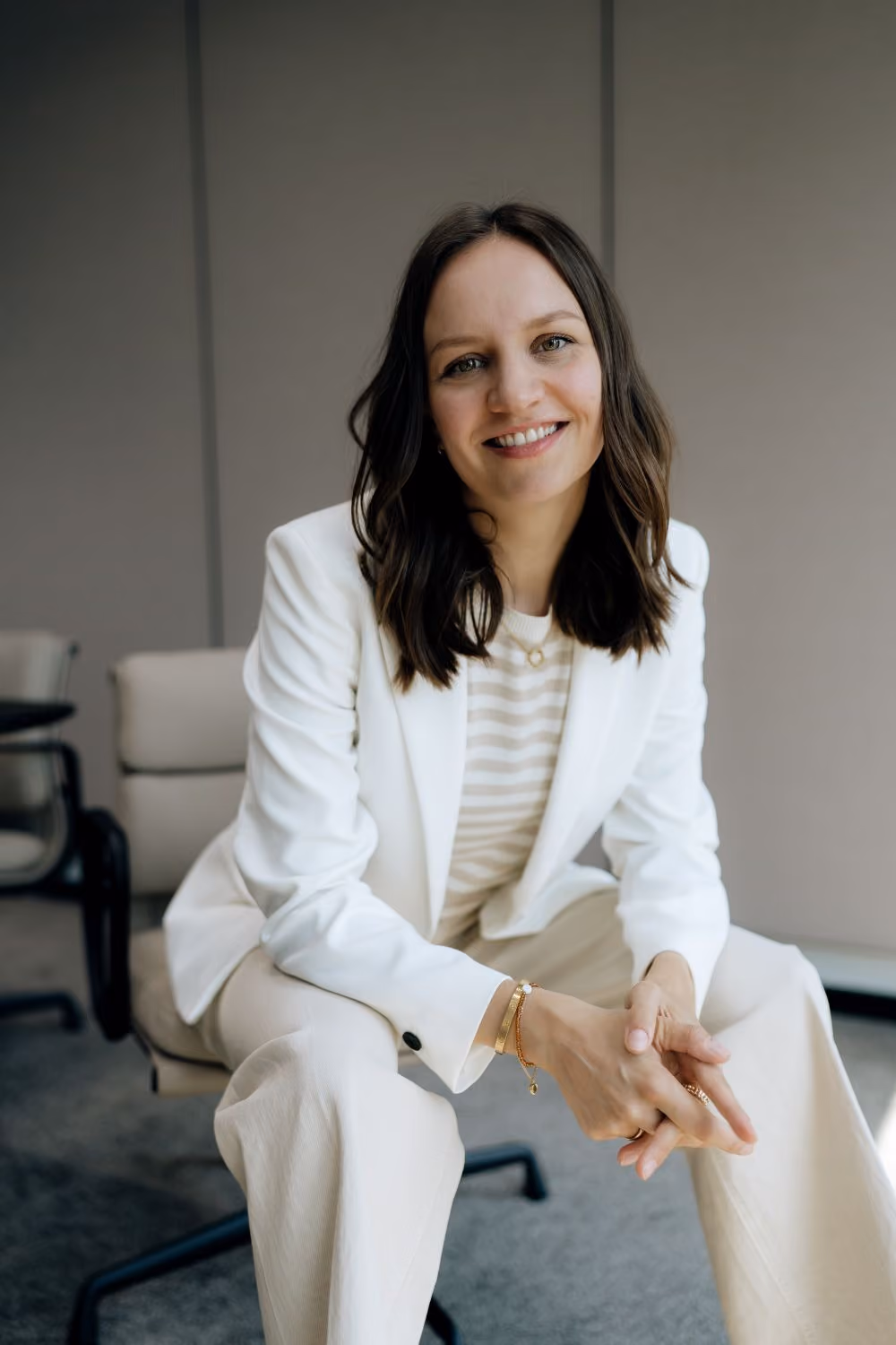Smiling woman with shoulder-length dark hair wearing a white blazer and cream pants sitting on an office chair with hands clasped.