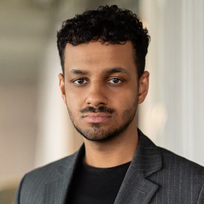 Headshot of a young man with short curly hair and a trimmed beard wearing a dark blazer and black shirt.