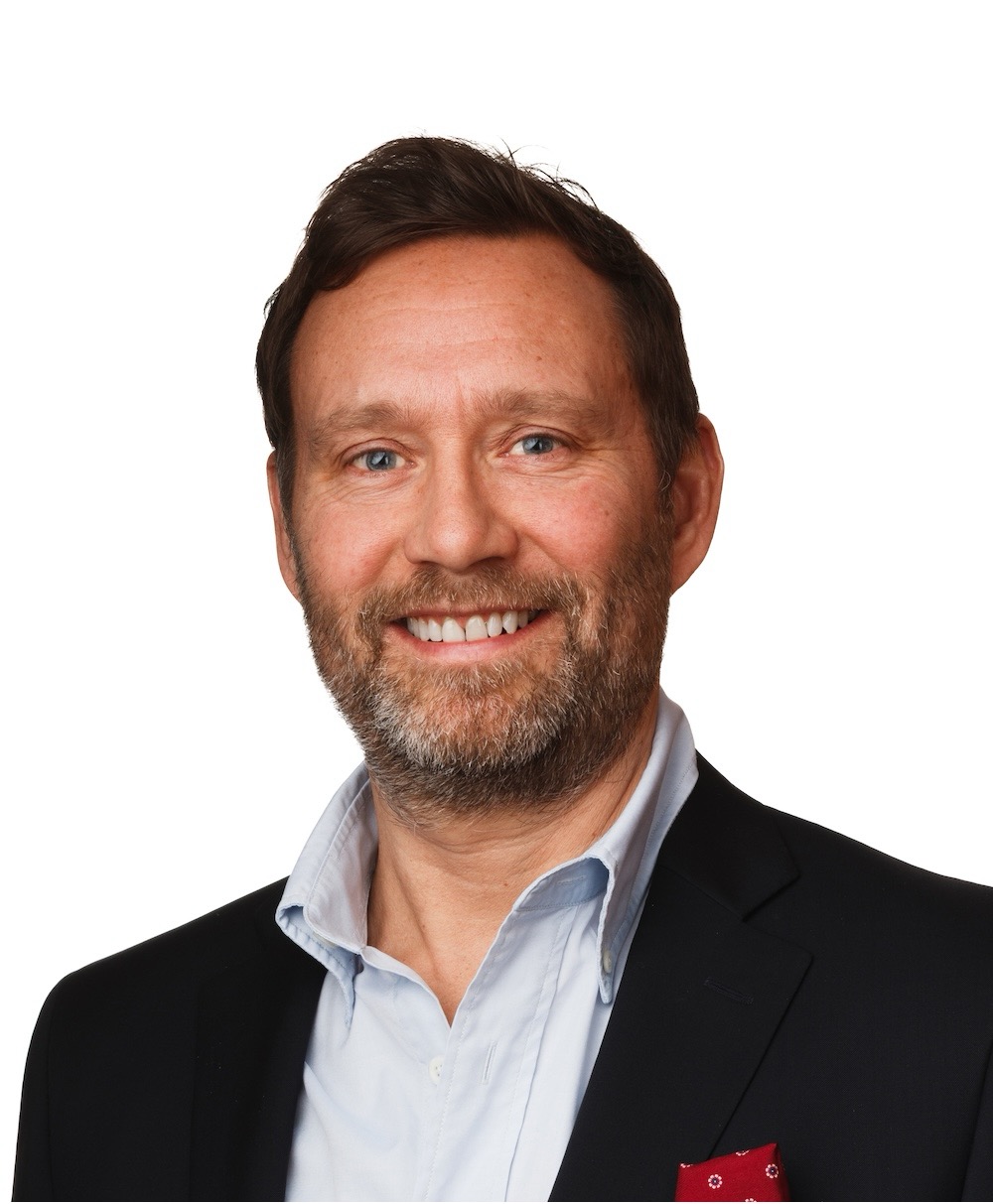 Smiling man with beard in a black jacket and light blue shirt against a white background.