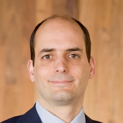 Headshot of a man with a receding hairline wearing a suit, smiling slightly against a wooden background.
