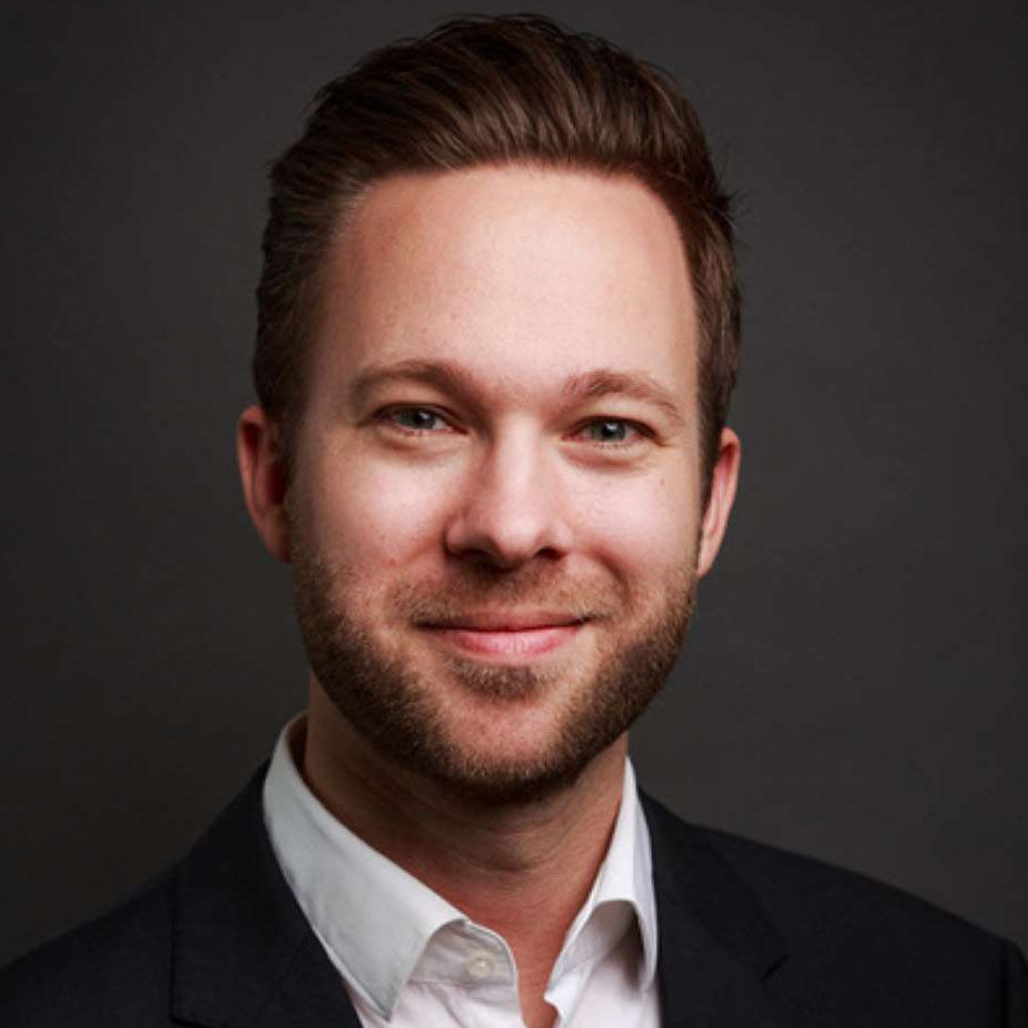 Smiling man with short brown hair and beard wearing a dark suit and white shirt against a dark background.