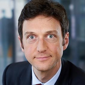 Man with short dark hair and blue eyes wearing a dark suit and white shirt, facing forward.