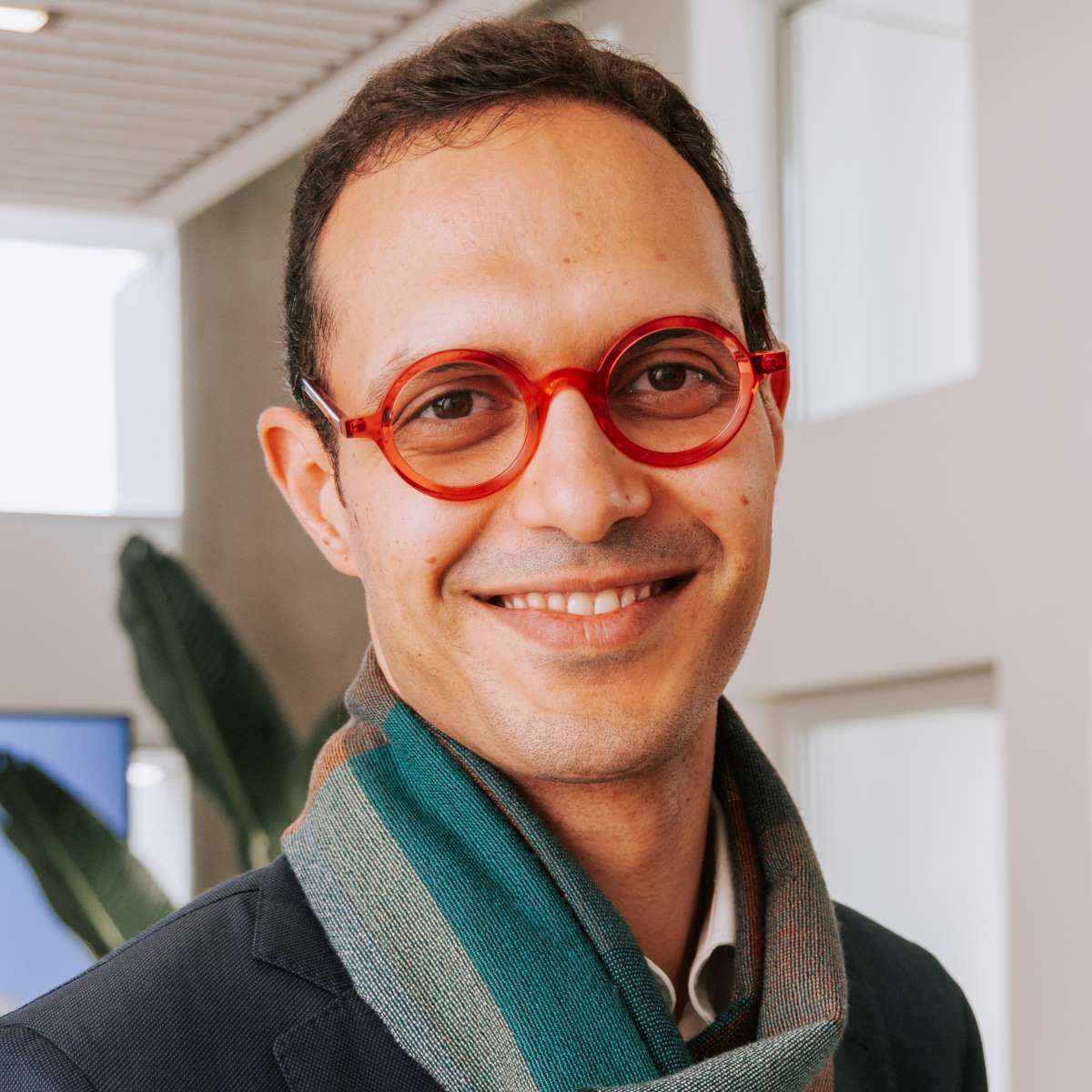 Smiling man wearing round red glasses and a green scarf in a bright indoor setting.