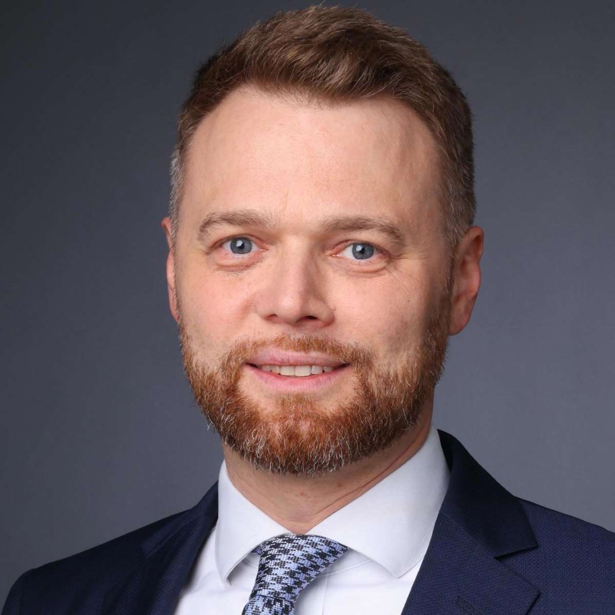 Smiling man with short brown hair and beard wearing a navy suit, white shirt, and patterned tie.