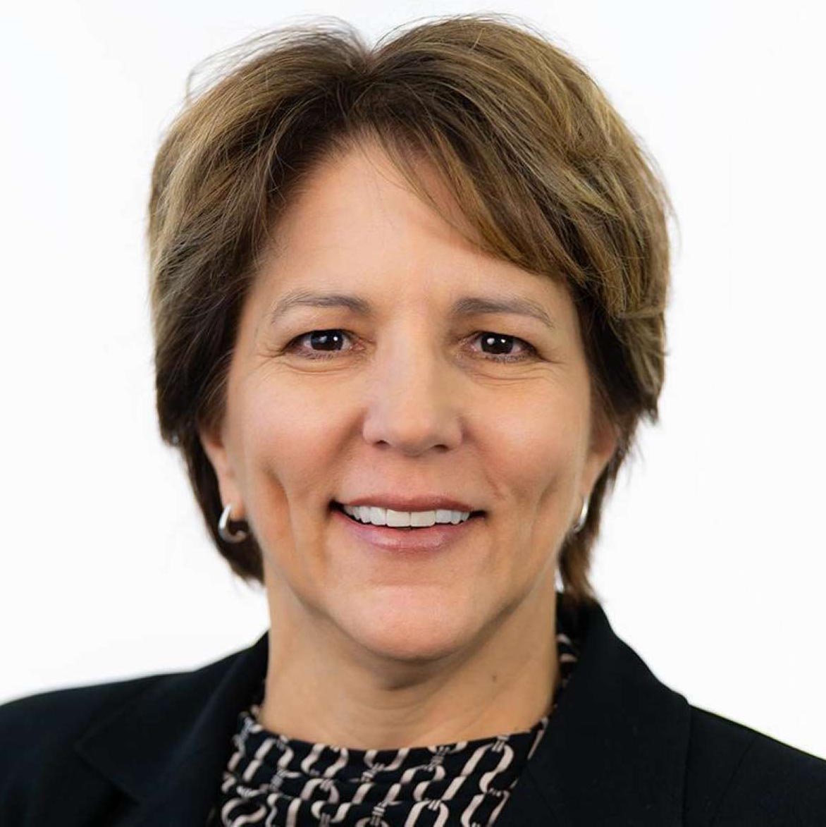 Smiling middle-aged woman with short brown hair wearing hoop earrings and a patterned blouse under a black jacket.