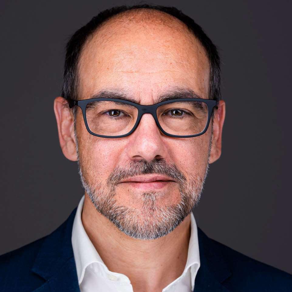 Middle-aged man with salt-and-pepper beard and glasses wearing a white shirt and dark jacket.