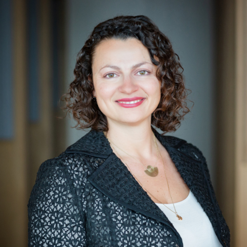 Smiling woman with curly hair wearing a black textured jacket and white top with gold necklaces.