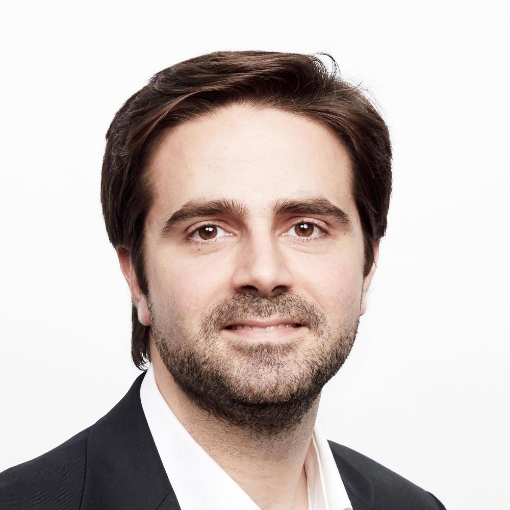 Portrait of a man with brown hair and beard wearing a black blazer and white shirt.