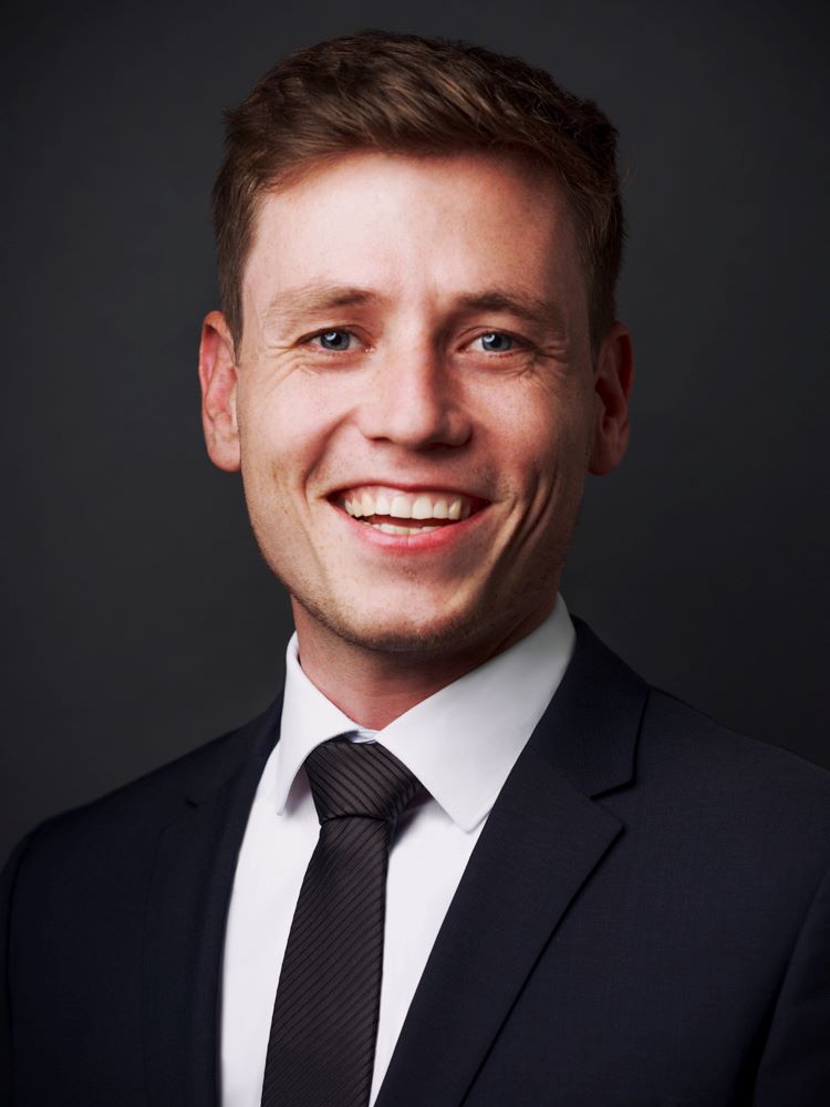 Smiling young man with short brown hair wearing a dark suit, white shirt, and black tie against a dark background.