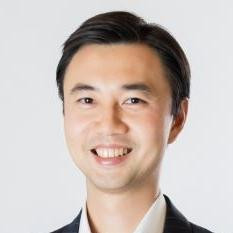 Portrait of a smiling man with dark hair wearing a dark suit and white shirt against a plain background.
