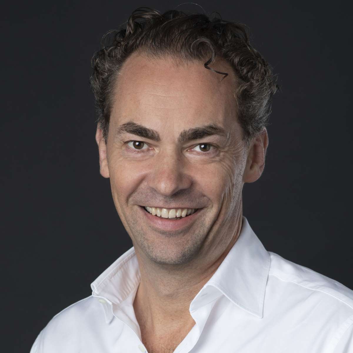 Smiling man with curly dark hair wearing a white shirt against a dark background.