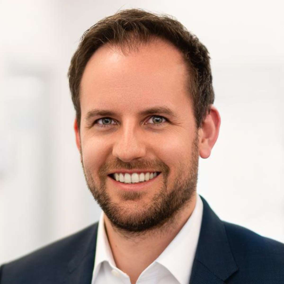 Smiling man with beard wearing a white shirt and dark blazer against a blurred background.