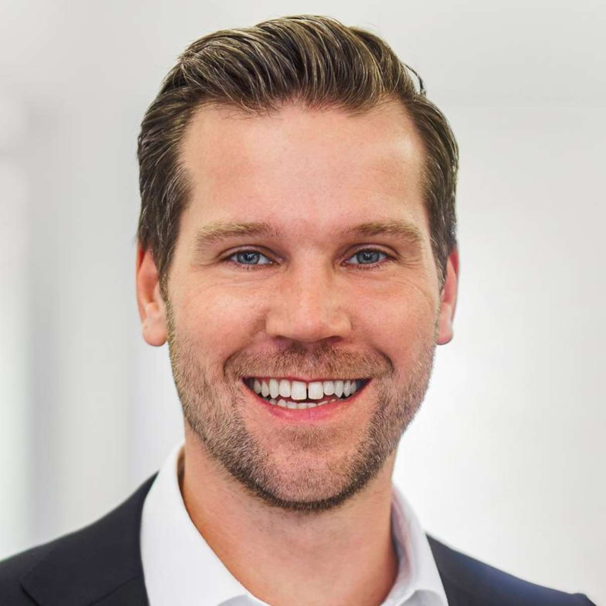 Smiling man with short brown hair and beard wearing a white shirt and dark blazer.