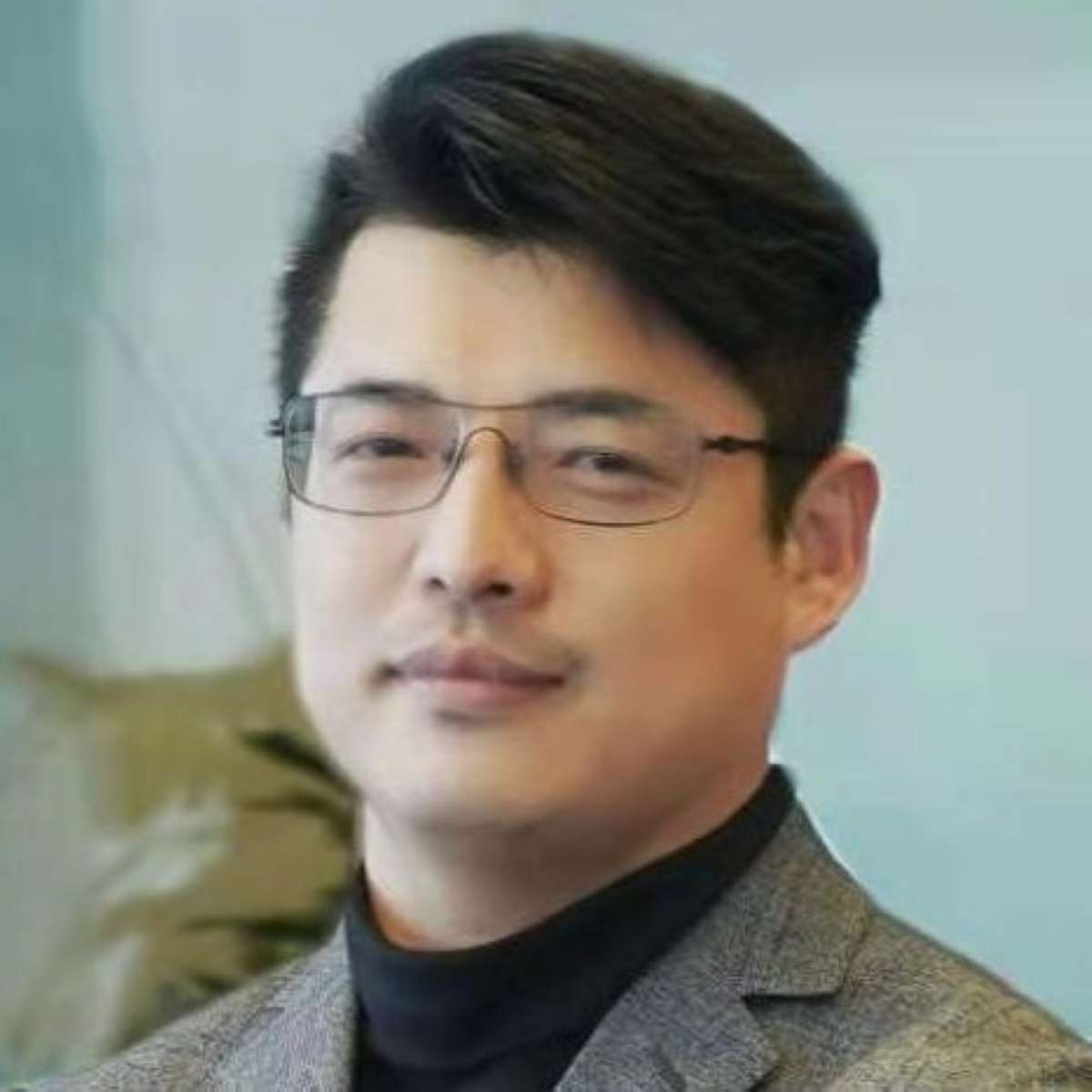 Man with glasses wearing a gray blazer and black turtleneck against a plain background.
