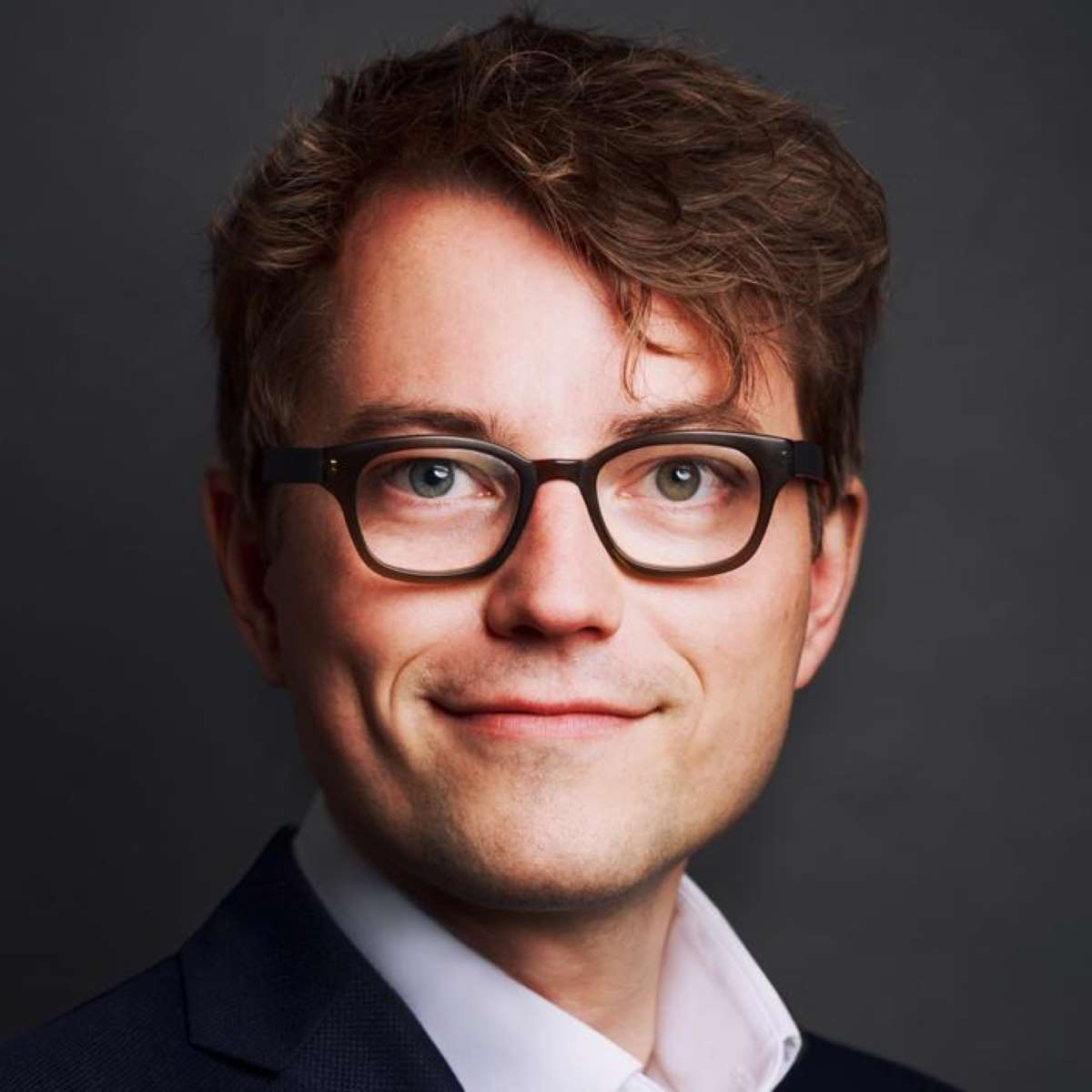Man with light skin, curly brown hair, glasses, and a slight smile in front of a dark background.