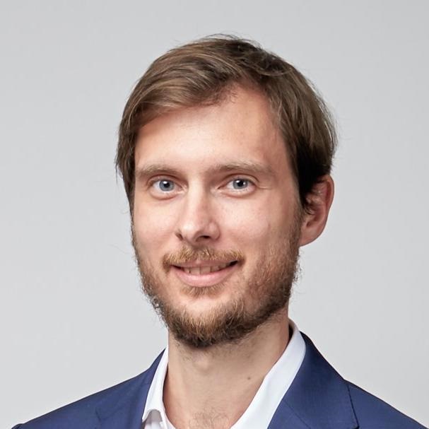 Young man with light brown hair and beard wearing a blue suit jacket and white shirt against gray background.