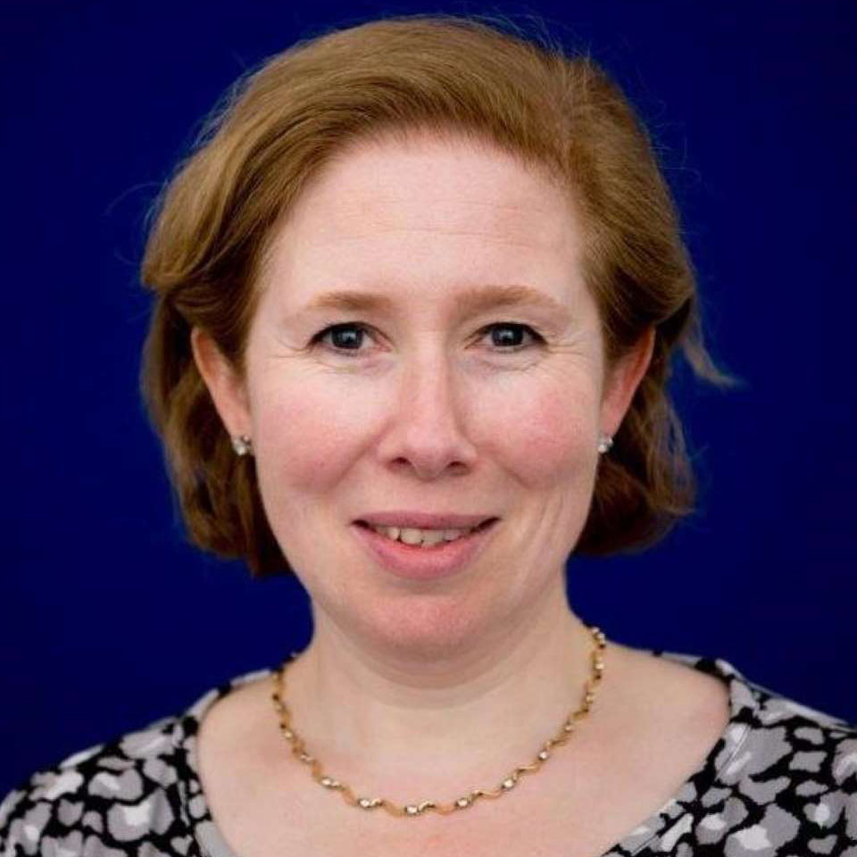 Smiling woman with short light brown hair, wearing a gold necklace and patterned top, blue background.
