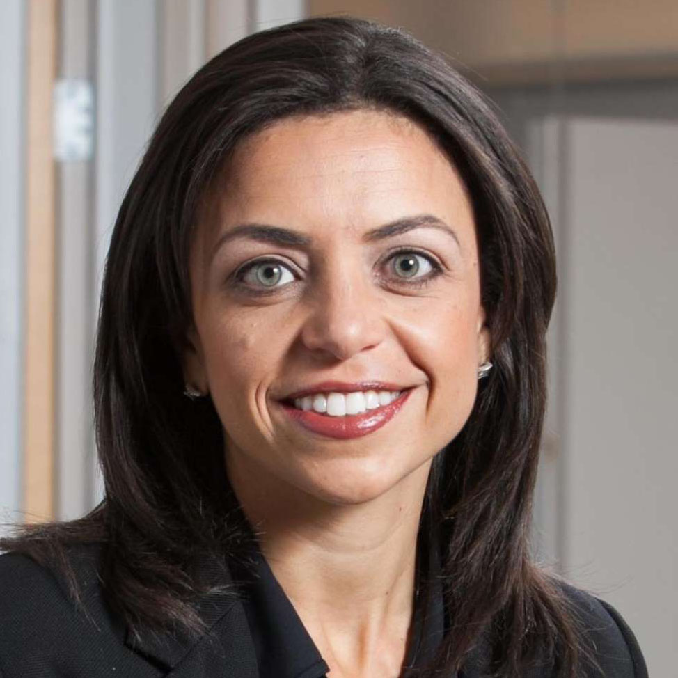 Smiling woman with dark hair and green eyes wearing a black blazer in an indoor setting.