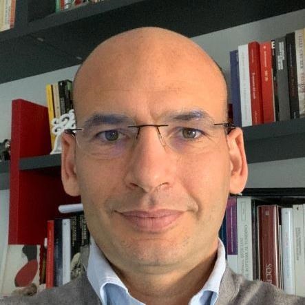 Bald man wearing glasses and a collared shirt stands in front of bookshelves with books.