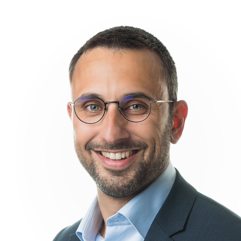 Smiling man with short hair, beard, glasses, blue shirt, and dark blazer against a white background.