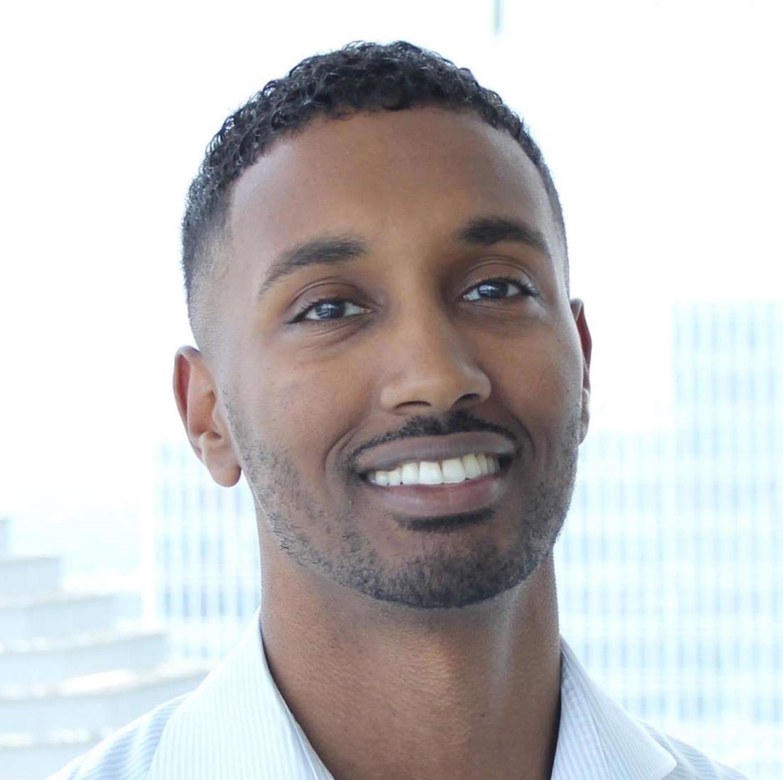 Smiling man with short curly hair and trimmed beard in a white shirt against a bright window.
