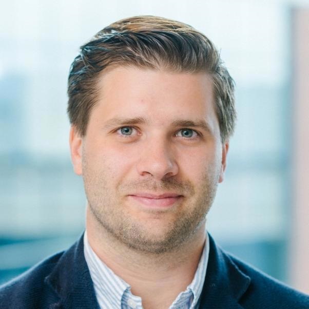 Portrait of a man with light brown hair and blue eyes wearing a navy jacket and a light blue shirt.