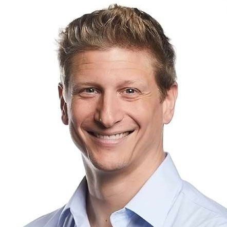 Smiling man with short wavy blond hair wearing a light blue collared shirt, against a white background.