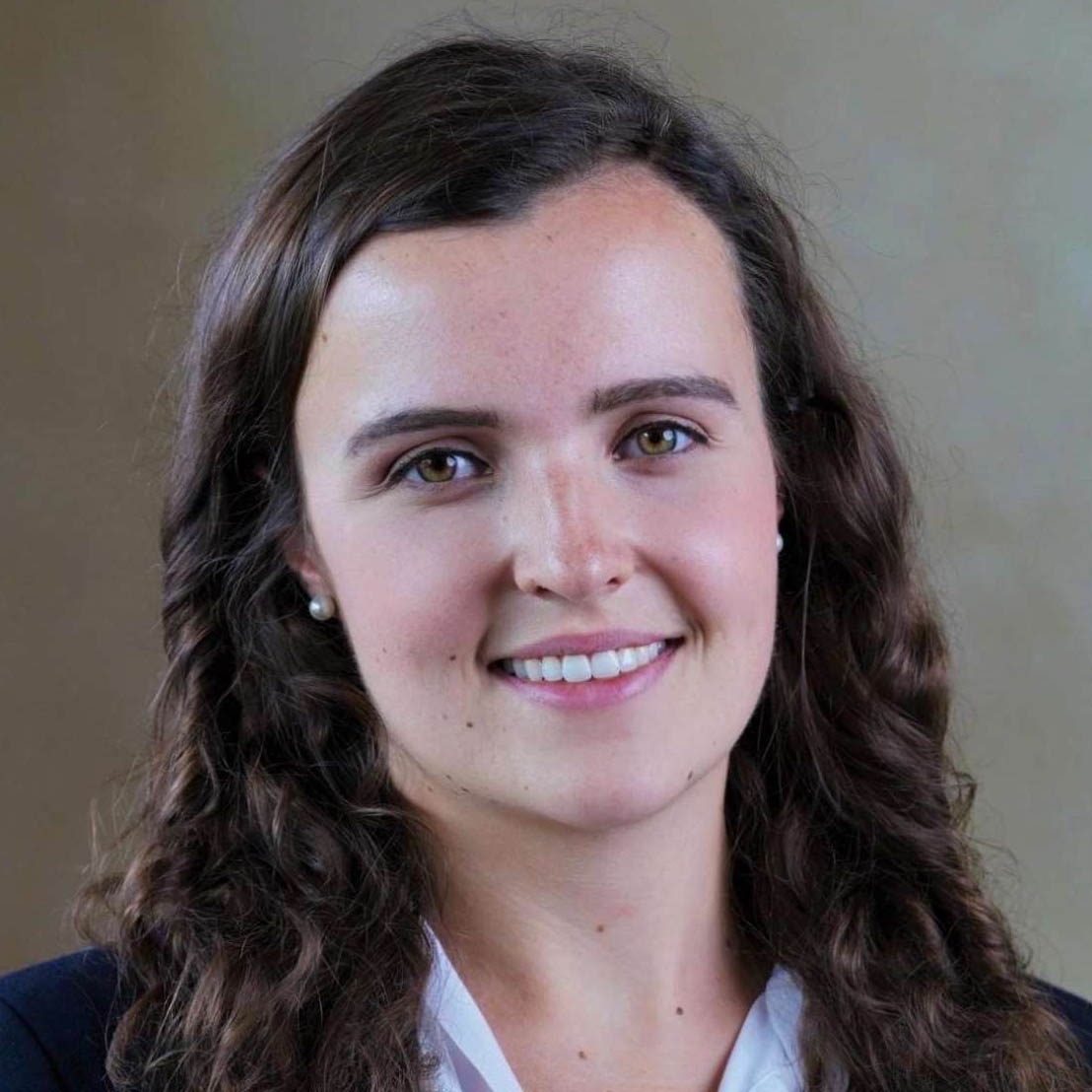 Smiling young woman with long curly brown hair and pearl earrings, wearing a white blouse and dark blazer.