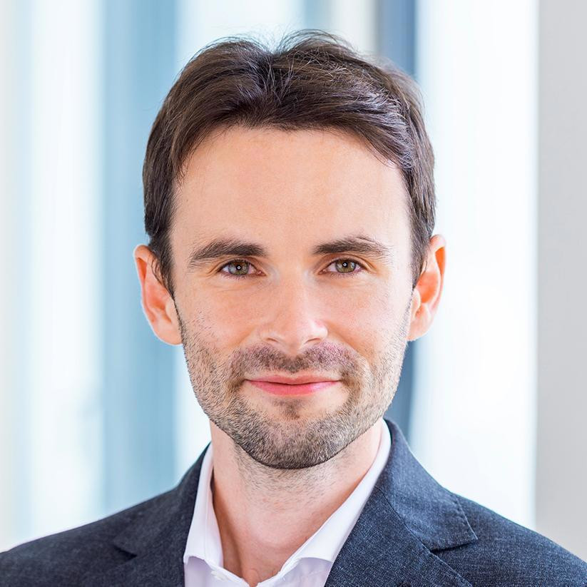 Portrait of a man with short brown hair, light beard, wearing a white shirt and dark blazer.