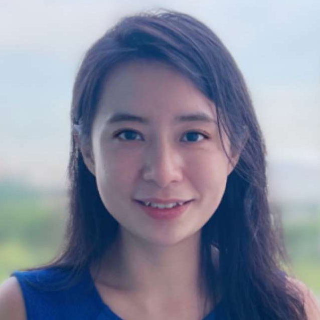 Portrait of a smiling woman with long dark hair wearing a blue top against a blurred outdoor background.