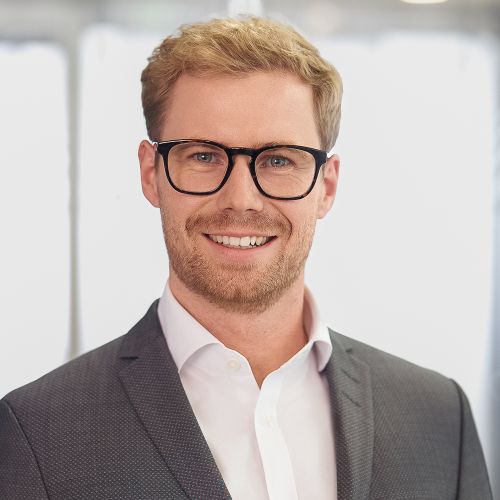 Smiling man with glasses, short blond hair, beard, wearing gray suit jacket and white shirt.