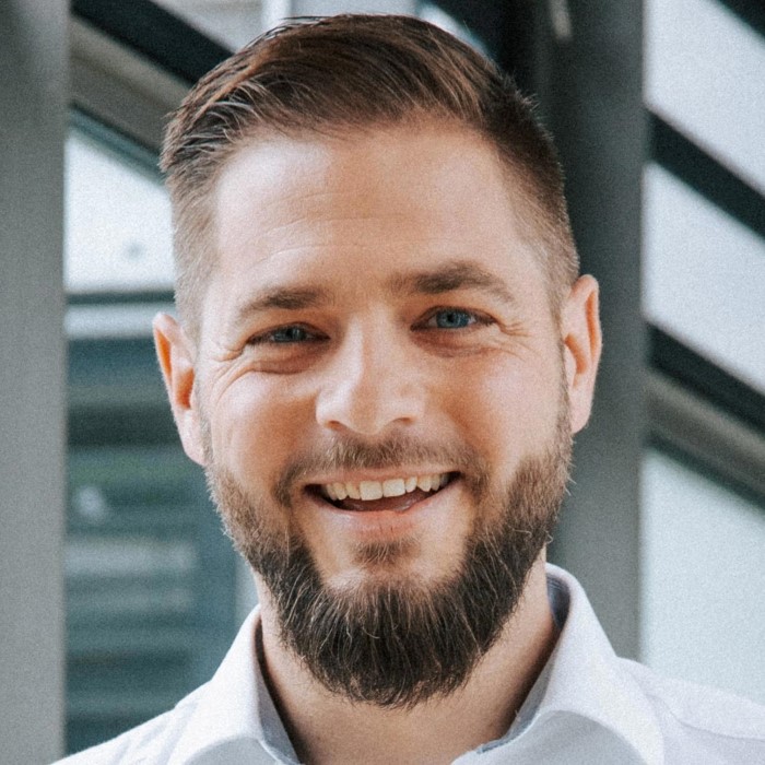 Smiling man with short brown hair and beard wearing a white collared shirt indoors.