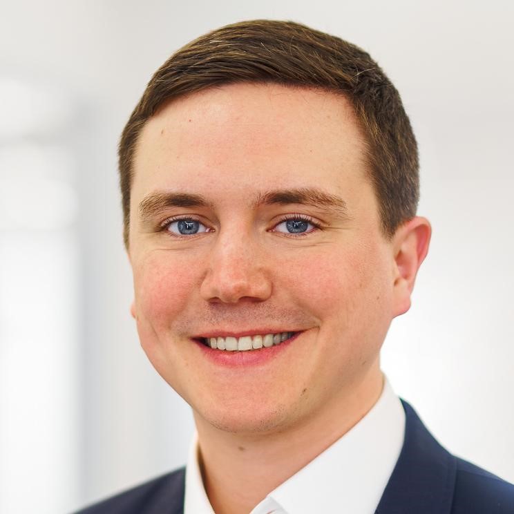 Smiling young man with short brown hair and blue eyes wearing a dark suit jacket and white shirt.