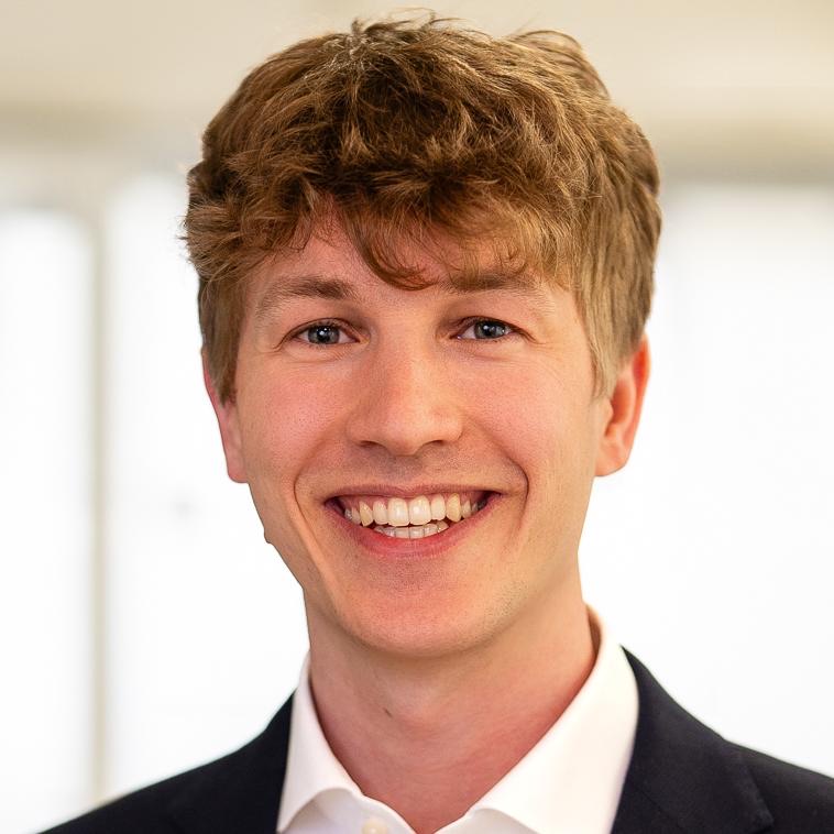Smiling young man with light brown hair in a suit jacket and white shirt against a bright background.