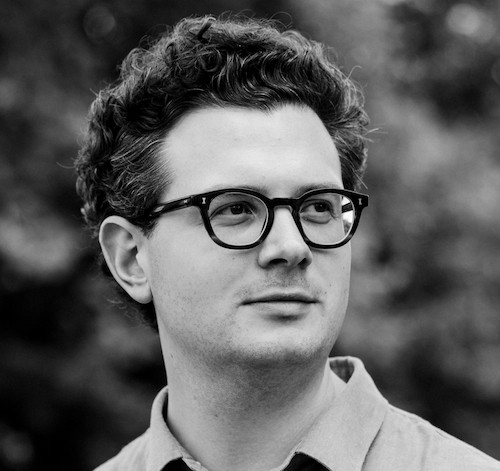 Black and white portrait of a man with curly hair and glasses looking slightly to the side outdoors.