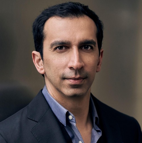 Portrait of a man with short black hair wearing a blue shirt and dark blazer against a blurred background.