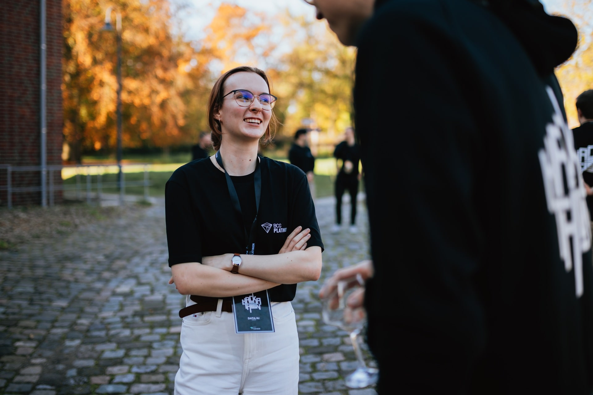 Young woman wearing glasses and a BCG Platinion shirt smiling outdoors with autumn trees in background.
