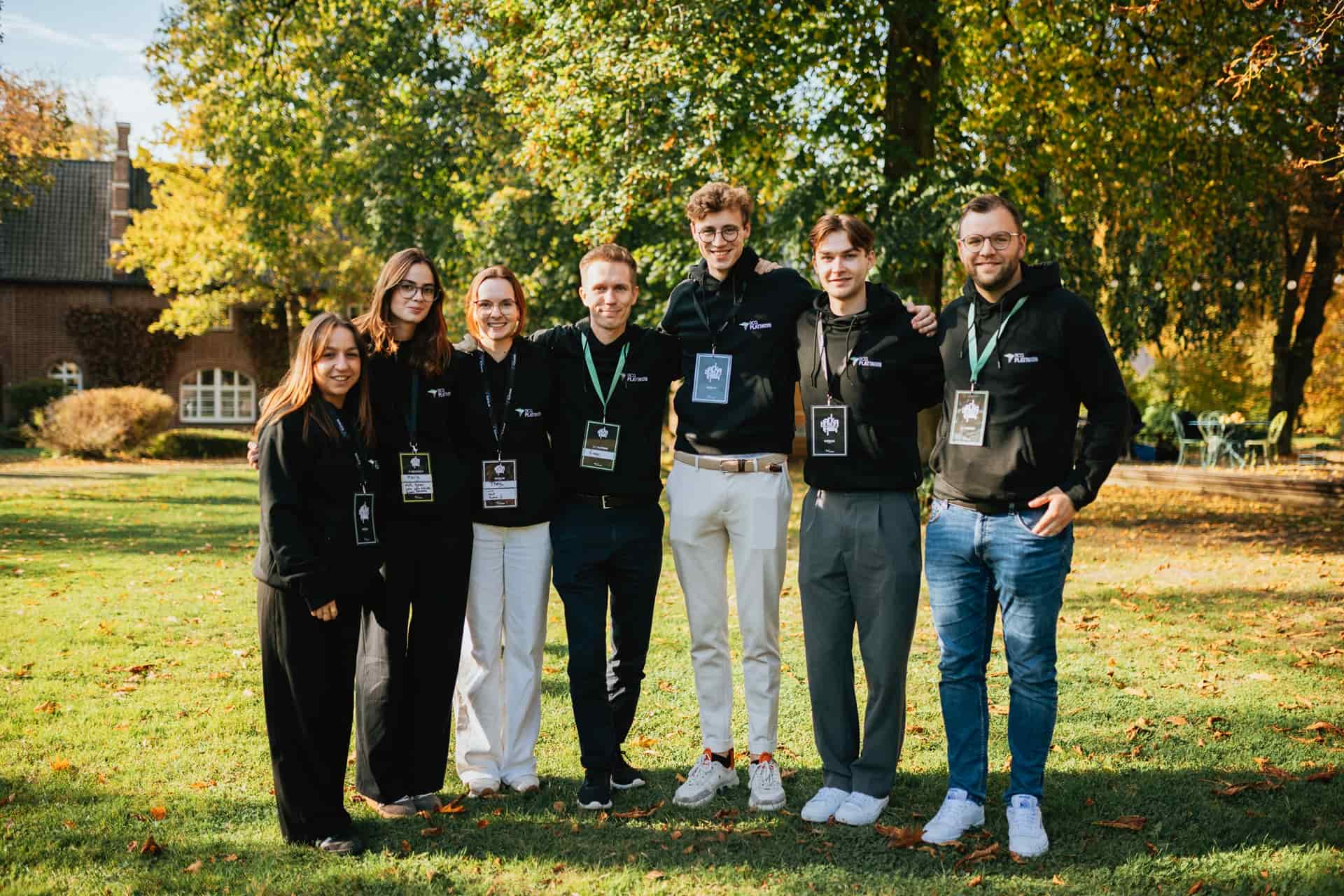 Seven people standing outdoors on grass wearing black hoodies and event badges, smiling with trees in background.