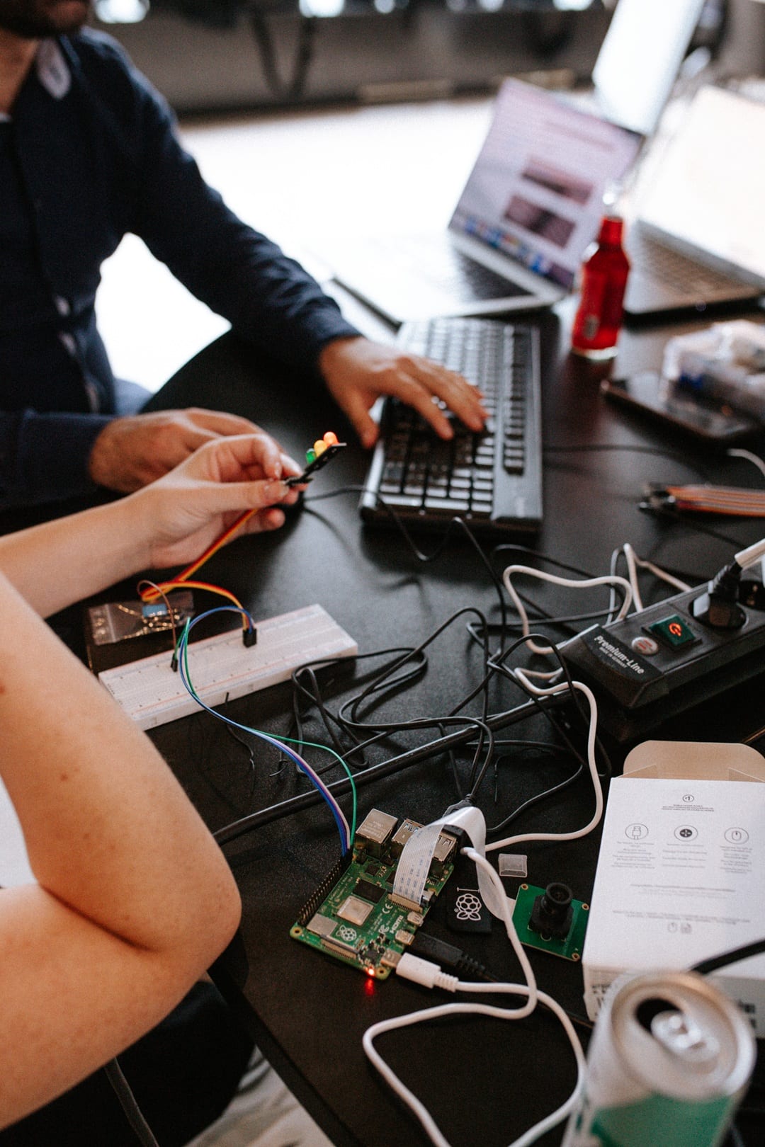 Two people working on electronics with a Raspberry Pi, LED lights, and laptops on a black desk.