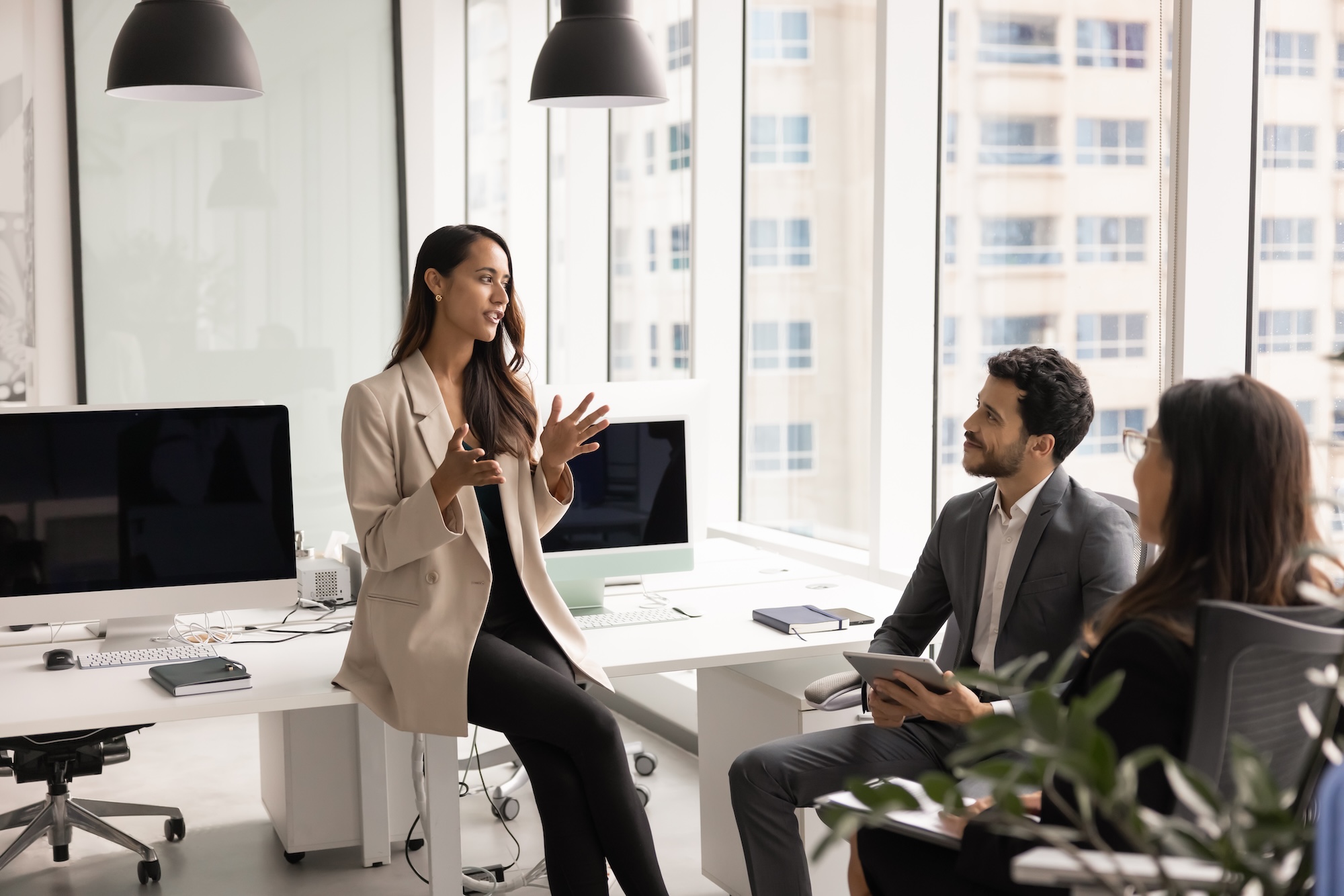Woman in beige blazer speaking to two seated coworkers in a modern office with large windows.